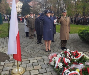 na zdjęciu wiele osób, na pierwszym planie wieńce oraz polska flaga, przed wieńcami dwie umundurowane osoby - policjantka i żołnierz salutują