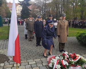 na zdjęciu wiele osób, na pierwszym planie wieńce oraz polska flaga, przed wieńcami dwie umundurowane osoby -  żołnierz i policjantka składająca kwiaty