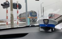 Czapka policjanta ruchu drogowego w radiowozie. W tle przejazd kolejowy oraz jadący pociąg i stojący przy przejeździe samochód ciężarowy.