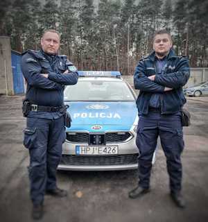 na zdjęciu dwóch umundurowanych policjantów, za nimi radiowóz