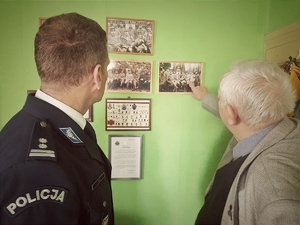 na zdjęciu w pomieszczeniu starszy mężczyzna pokazuje palcem zdjęcie wiszące na ścianie, obok mężczyzny stoi policjant