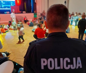 Zdjęcie przedstawia tył umundurowanego policjanta w tle bawiące się dzieci.