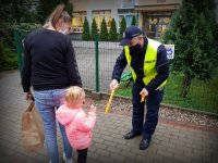 Policjant pod przedszkolem wręcza odblask dziewczynce, która trzyma mamę ze rękę