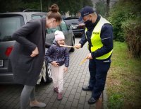 Policjant na chodniku wręcza odblask uśmiechniętej dziewczynce obok której stoi kobieta