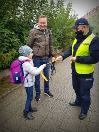 Policjant wręcza na chodniku odblask dziewczynce koło której stoi mężczyzna.