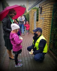 Policjant kuce przed dziewczynką i wręcza jej odblaski. Obok dziecka stoi kobieta która ma nad głową otwarty parasol.